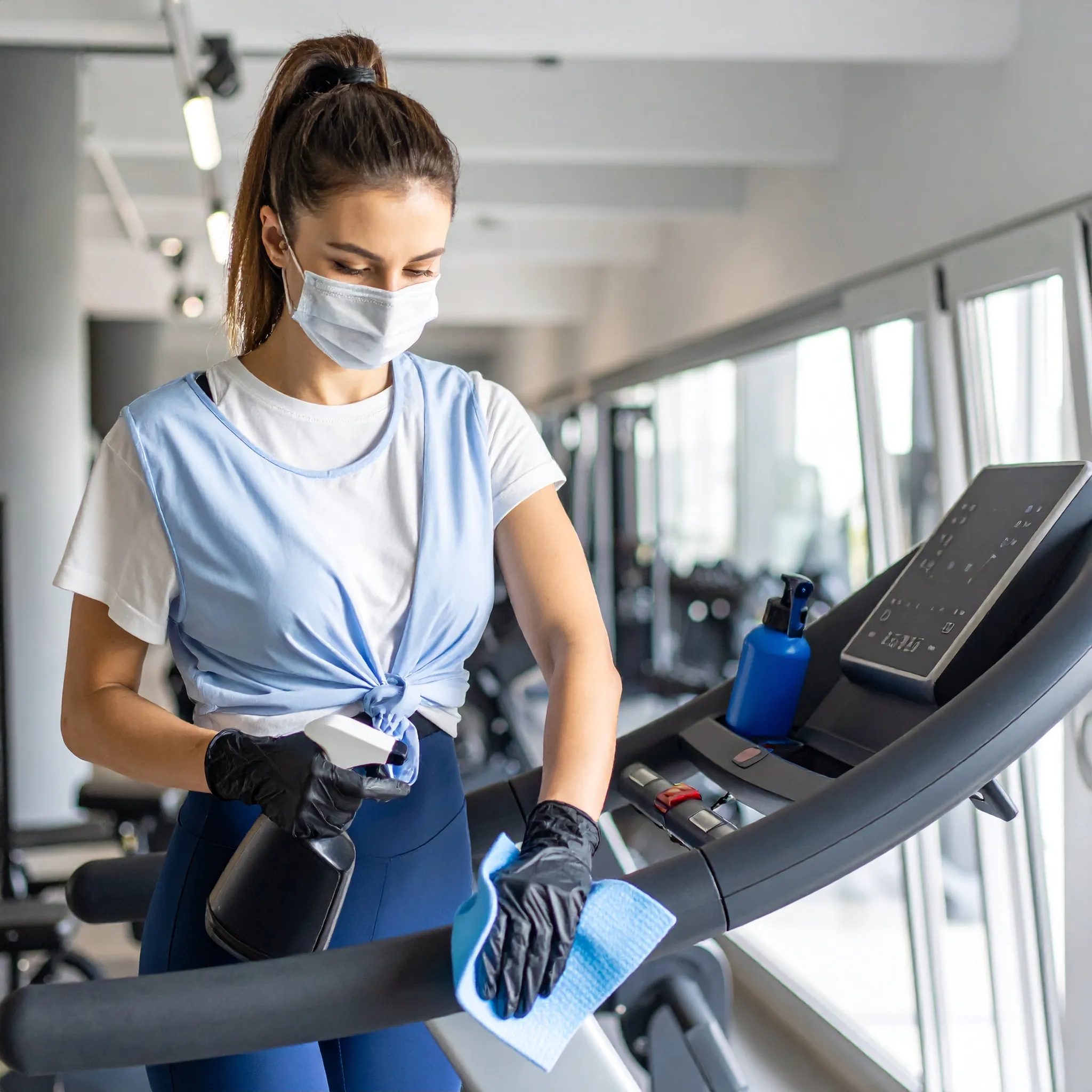 Reinigungskraft mit Maske desinfiziert ein Laufband im Fitnessstudio